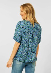 Blusa a maniche corte in tessuto blu con un motivo floreale verde e nero. Presenta un pannello posteriore e dettagli di piega per un maggiore comfort.