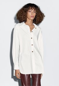 Bílá košile na knoflíky s límcem, dlouhými rukávy a hnědými knoflíky. Tkanina je hladká a košile má uvolněný střih. Kombinováno s lesklými vínovými kalhotami.