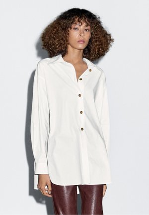 White button-up shirt featuring a collar, long sleeves, and brown buttons. The fabric appears smooth with a relaxed fit. Paired with shiny burgundy pants.