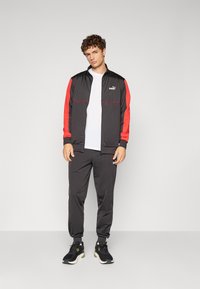 Sort sportjakke med røde accenter og Puma-logo, parret med matchende sorte bukser. Modellen har sorte sneakers på. Glat stoftekstur.