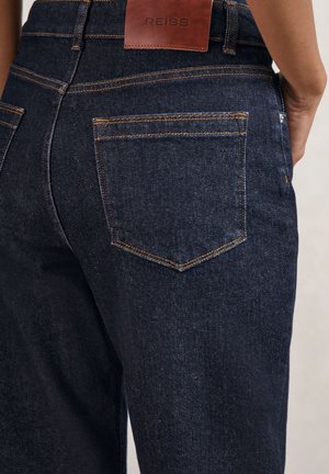 Tummansiniset denimhousut oransseilla saumoilla ja ruskealla Reiss-merkkisellä nahkalapulla takavyötäröllä, yllään henkilö, joka näkyy vyötäröstä alaspäin.