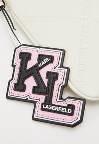 Černý a růžový gumový logo štítek s textem "KL" a "KARL LAGERFELD"; texturovaný povrch s bílým okrajem; připevněn na hladkém bílém pozadí.