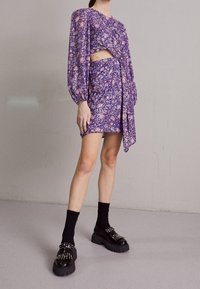 Fialové květinové šaty s dlouhými rukávy, výstřihem v pase, nabraným designem a postranní stahovací páskou. Doplněné robustními černými platformovými botami.