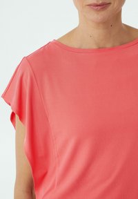 Koralowy różowy top, wykonany z gładkiego materiału, charakteryzuje się szerokim dekoltem i krótkimi, falbaniastymi rękawami oraz luźnym krojem. Widoczna delikatna faktura.