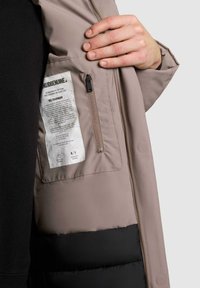 Giubbotto trapuntato beige con la parte inferiore nera, dotato di tasca interna con zip e etichetta per la cura. Tessuto liscio con una forma strutturata.