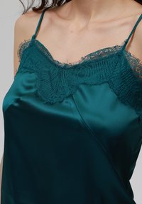 Teal satengcamisole med delikat blondertrim ved utringningen, med tynne stropper og en glatt, glansfull tekstur.