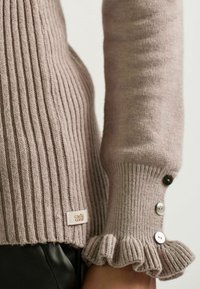 Close-up van een persoon die een taupe geribbelde trui draagt met gefronte mouwen, voorzien van drie knopen op de mouw en een klein stoffen label.