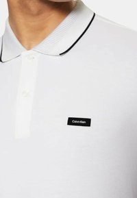 Polo bianco con bordo del colletto nero e patta con due bottoni. Presenta un piccolo logo nero di Calvin Klein sul petto sinistro. Tessuto con texture.