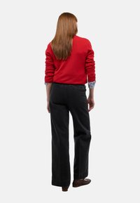 Pull en tricot rouge à manches longues, associé à un pantalon large noir taille haute. Le modèle se tient de dos par rapport à l'appareil photo.