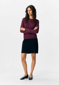 Un cardigan bourgogne avec des boutons, associé à une jupe noire avec une fente avant. Des formes simples et ajustées ainsi que des textures douces sur un fond neutre.