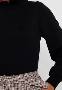 Černý pletený top s volánkovým stojáčkem a balónovými rukávy, doplněný sukní s kostkovaným vzorem a uvázaným opaskem.