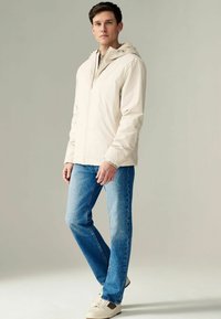 Jachetă impermeabilă bej cu glugă, fermoar frontal și două buzunare laterale, asociată cu blugi din denim albastru și pantofi bej.