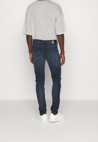 Hombre con jeans slim de color azul oscuro, zapatillas blancas y una camiseta de rayas en gris y blanco, de pie frente a un fondo blanco liso.