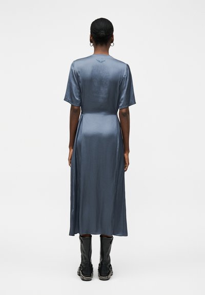 Γυναίκα με σκούρα μαλλιά σε κότσο, που φοράει μπλε σατέν μεσαίο φόρεμα και μαύρα μποτάκια αστραγάλου, στέκεται γυρισμένη πλάτη σε λευκό φόντο.