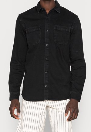 Chemise noire en coton à manches longues avec un devant à boutons, deux poches poitrine et un col classique. Le tissu a une finition légèrement texturée.