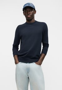 Μπλε πλεκτό πουλόβερ με στρογγυλή λαιμόκοψη και ριμπ μανσέτες, σε συνδυασμό με ανοιχτόχρωμο τζιν παντελόνι και καπέλο με μπλε μοτίβο.