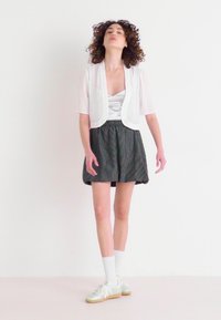 Biela polopriehľadná bunda s krátkym rukávom cez strieborný top a čierne pásikované šortky. Biele podkolienky a svetlozelené tenisky dotvárajú outfit.