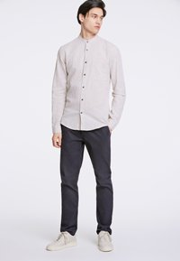 Camisa abotonada gris claro con cuello mao, que cuenta con un bolsillo frontal y botones más oscuros, combinada con pantalones marinos y zapatillas blancas.