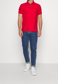 Červené polo tričko se dvěma knoflíky u límce, krátké rukávy a malé logo na hrudi. V kombinaci s modrými džíny a bílými teniskami.