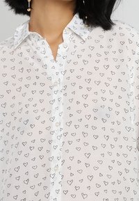 Chemise blanche boutonnée avec de petits contours de cœur noirs portée par une personne aux cheveux noirs et des boucles d'oreilles gouttes en or.