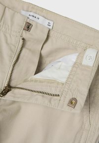 Beżowe spodnie z zamkiem błyskawicznym w odcieniu złota, z zapięciem na guzik i wewnętrzną kieszenią. Metka jest widoczna wewnątrz pasa.