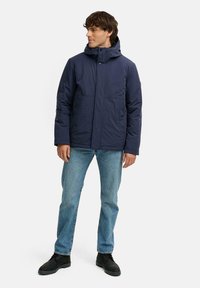 Marineblå pufferjakke med hætte, der har en forreste lynlås, trykknapper og to sidelommer. Båret over lyseblå jeans og sorte støvler.