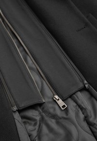 Svart ytterjacka med dubbel dragkedja, texturerad utsida och slät innerfoder. Har ett framträdande varumärkesmärke på dragkedjans draghandtag.