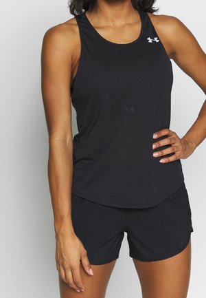 Femme portant un haut de sport noir sans manches et un short noir, debout avec une main sur la hanche devant un fond uni.