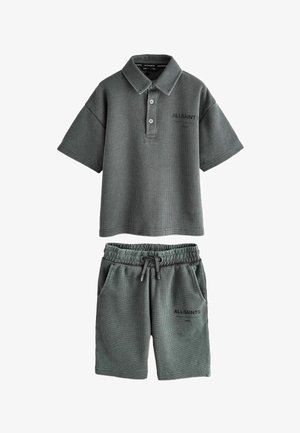 Teenager-Junge steht und trägt ein graues, übergroßes Poloshirt sowie passende knielange Shorts, weiße Sneakers und Socken mit Logo vor einem einfarbigen Hintergrund.