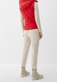 Gilet matelassé rouge avec capuche, associé à un pantalon de jogging beige et des bottes de combat beige clair. Textures lisses et design décontracté.