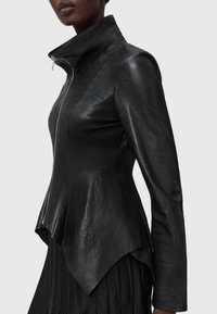 Schwarze Lederjacke mit hohem Kragen, strukturiertem Design, asymmetrischem Saum und Reißverschluss. Glatte Textur mit glänzenden Akzenten.