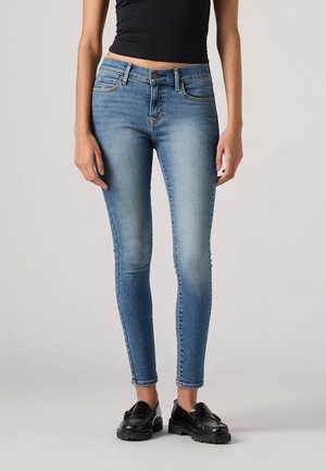Jean bleu slim fit avec devant délavé porté avec des mocassins noirs et un top noir court sur une personne debout devant un fond uni.
