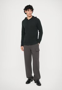 Haut à capuche en tricot vert bouteille avec un motif en câble, doté d'une poche avant et de cordons de serrage, associé à un pantalon cargo large gris et des chaussures noires.