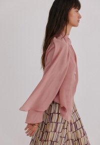 Vrouw in een vloeiende roze blouse met cape-mouwen en een gestreepte rok die zijwaarts loopt tegen een eenvoudige lichte achtergrond.