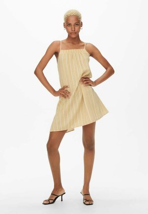 Robe jaune à rayures avec des bretelles fines, un décolleté froncé, une coupe décontractée et un ourlet asymétrique. Portée avec des sandales noires à lanières.