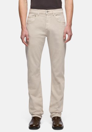 Beige bukser med lige ben, båret af en person, der står med afslappede hænder, kombineret med mørkebrune lædersko og en sort skjorte.