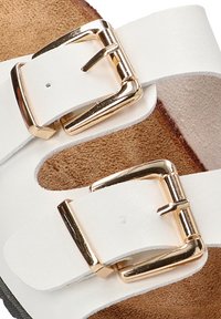 Correa de sandalia de cuero blanco con hebillas de metal dorado, que presenta una plantilla interior texturizada en tono marrón. Enfoque en los detalles de la hebilla y el diseño de la correa.