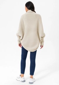 Beige oversized gebreide trui met een ribbelstructuur, voorzien van een sjawlzoom en losse mouwen, gecombineerd met donkerblauwe spijkerbroeken en witte sneakers.