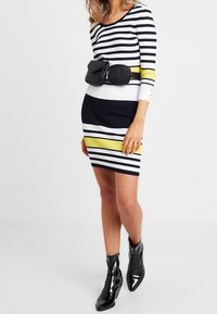 Robe en tricot à rayures horizontales bleu marine, blanche et jaune, avec manches longues. Associée à une ceinture-sac noire et des bottines noires brillantes.
