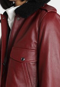 Röd läderjacka med en svart fårskinnskrage, dragkedjestängning och en framficka med en svart tryckknapp. Slät textur.