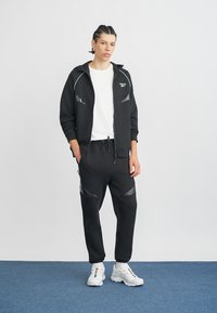 Melns sporta tērps ar rāvējslēdzēju jakas augšdaļā un biksēm ar reljefiem akcentiem. Balta T-krekls zem tā, kombinēts ar masīvām baltām sporta kurpēm.