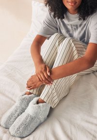 Chaussons en fausse fourrure gris clair avec un design arrondi, une texture douce et un bord en peluche, portés avec un pantalon à rayures de couleur claire sur un lit.