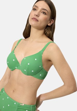 Mujer con un sujetador verde texturizado con lunares blancos y pantalones de talle alto a juego, mirando ligeramente hacia arriba y hacia un lado.