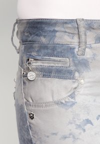 Jeans de mezclilla de color azul claro con un patrón desvaído, diseño de cinco bolsillos, herrajes plateados y un pequeño charm adjunto al bolsillo delantero.