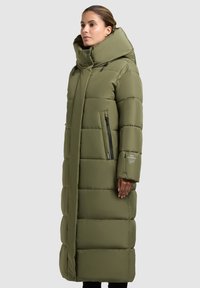 Garšs olīvzaļš pūkains mētelis ar augstu apkakli, ar rāvējslēdzēju sānu kabatām un šūtu, kviltētu dizainu. Gluda tekstūra ar minimālistisku izskatu.