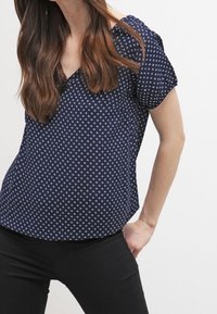 Blusa blu navy a maniche corte con scollatura a V, caratterizzata da un motivo geometrico bianco. Tessuto morbido con una vestibilità comoda e orlo arrotondato.