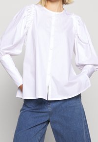 Camicia bianca con maniche a sbuffo, caratterizzata da tessuto arricciato sulle spalle. Texture liscia, vestibilità rilassata e orlo dritto.