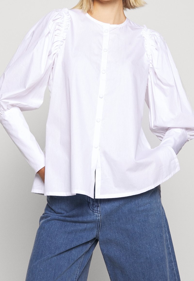 Camicia bianca con maniche a sbuffo, caratterizzata da tessuto arricciato sulle spalle. Texture liscia, vestibilità rilassata e orlo dritto.