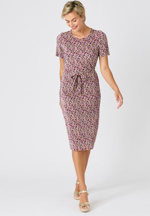 Mujer con vestido floral de manga corta y largo hasta la rodilla, con un lazo en la cintura, caminando y mirando hacia abajo, sonriendo sobre un fondo liso.