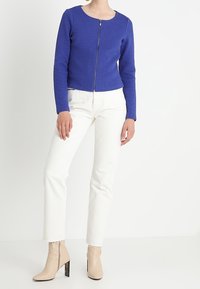 Blå texturerad zip-up jacka med rund halsringning, kombinerad med vita raka byxor och beige ankelskor med klack.